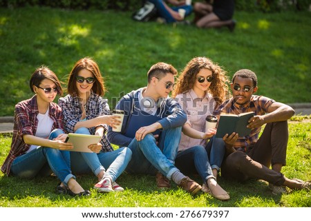 Group of young attractive smiling students dressed casual sitting on the lawn in park and studying.