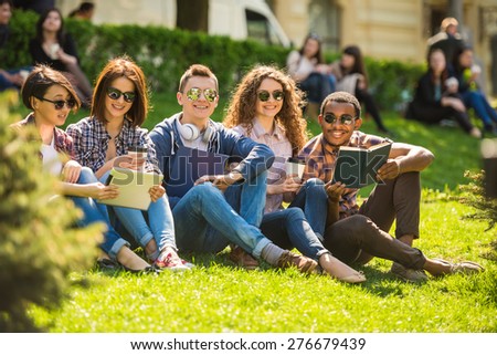 Group of young attractive smiling students dressed casual sitting on the lawn in park and studying.