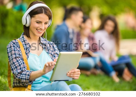 Young beautiful smiling student sitting on the lawn at park and listen to music.