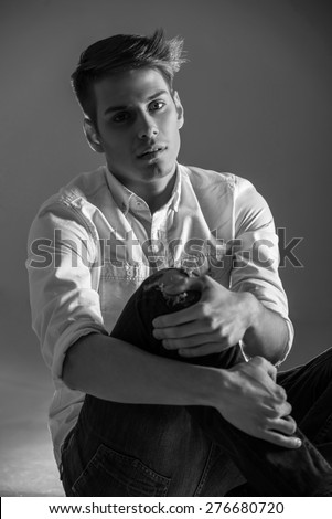 Young handsome man in shirt and jeans sitting on the floor. Black and white fashion portrait.