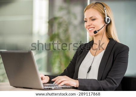 Beautiful young woman with headset sitting at office and using laptop.