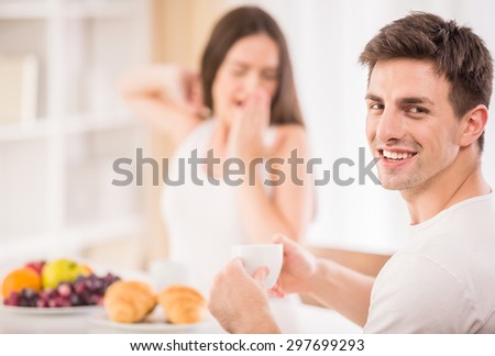 Lazy morning in the kitchen. Young attractive man drinking tea while his sleepy girlfriend making yawn.