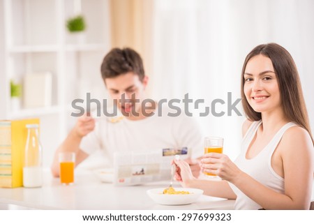 Young woman sitting at the table in the kitchen and eating while her boyfriend reading newspaper on background.