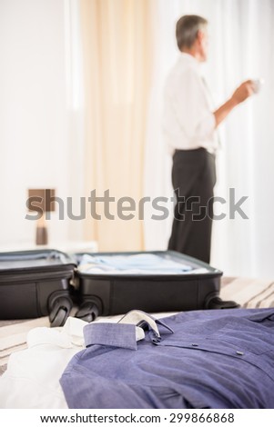 Suitcase and clothes on the bed in hotel room. Back view of man with cup of coffee looking at the window.