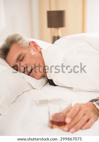Businessman sleeping on bed after hard working day with glass of whiskey and his phone at the hotel room. Side view.