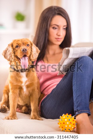 Picture of young woman is reading a newspaper while her dog is sitting on sofa near her.