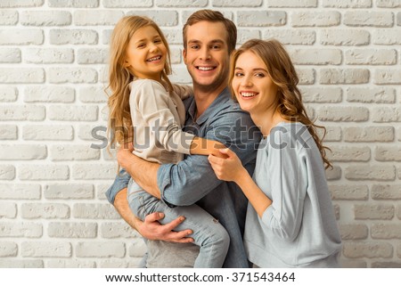 Beautiful young family hugging, looking in camera and smiling while standing against white brick wall