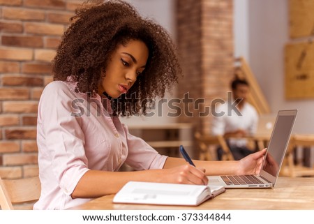 Young beautiful Afro-American businesswoman using laptop and writing in notebook while studying in cafe
