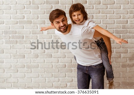 Portrait of a handsome father carrying his cute son on back. Both in white t-shirts smiling, standing against white brick wall.