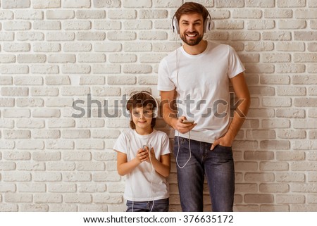Handsome young father and his cute little son using smart phones, listening to music, looking in camera and smiling. Both in white t-shirts and jeans, standing against white brick wall.