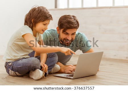 Handsome young father in casual clothes and his cute little son using a laptop while lying on a wooden floor in the room
