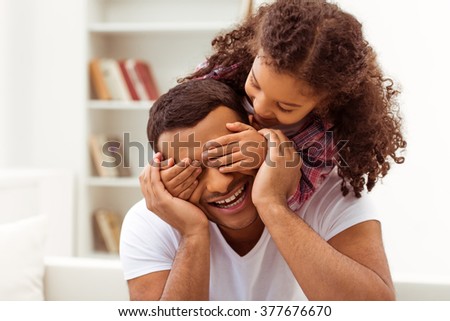 Cute little Afro-American girl in casual clothes covering her father eyes. Both smiling.