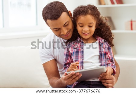 Cute little Afro-American girl in casual clothes sitting on her father\'s knees. Both using a tablet and smiling.