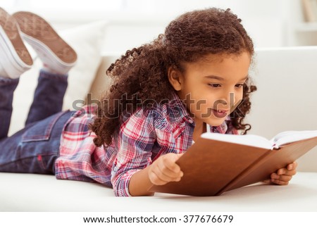 Cute little Afro-American girl in casual clothes reading a book and smiling while lying on a sofa in the room.
