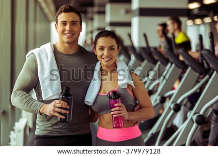 Attractive young people holding bottles of water, looking at camera and smiling while standing in gym