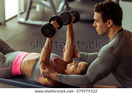 Attractive young woman working out with dumbbells in gym, handsome muscular trainer helping her