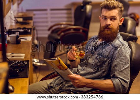Handsome young bearded man reading a magazine and holding a glass of beverage while sitting on a chair at the barber shop
