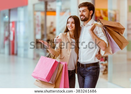 Happy beautiful young couple holding shopping bags, looking upon showcase and smiling while standing in mall