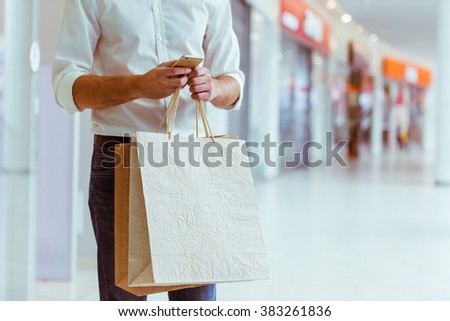 Handsome young man in white shirt holding shopping bags and using a smart phone while standing in mall, close-up