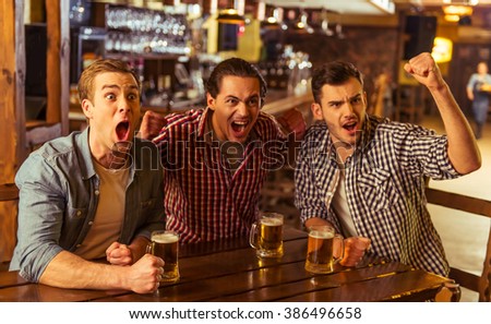 Three young men in casual clothes are cheering for football and holding glasses of beer while sitting in pub
