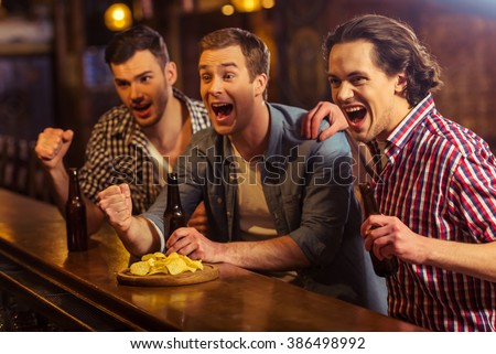 Three young men in casual clothes are cheering for football and holding bottles of beer while sitting at bar counter in pub