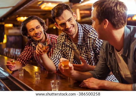 Three young men in casual clothes are talking, laughing and drinking while sitting at bar counter in pub