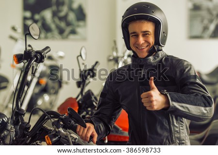 Attractive young blond man in black leather jacket and helmet is looking at camera, showing Ok sign and smiling while sitting on a motorbike