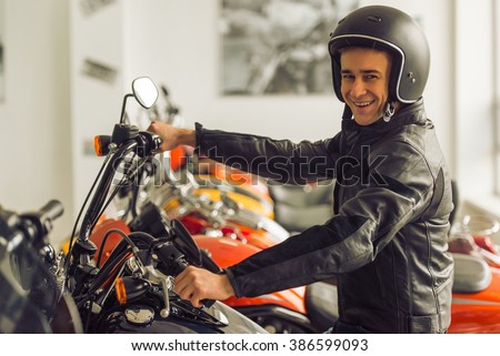 Side view of attractive young blond man in black leather jacket and helmet looking at camera and smiling while sitting on a motorbike