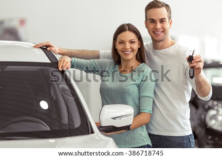 Beautiful young couple is smiling and looking at camera while leaning on their new car in a motor show. Man is holding car keys