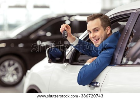 Handsome young businessman in classic blue suit is smiling, looking at camera and showing car keys while sitting in car in a motor show