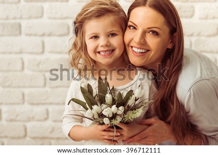 Beautiful young mother and her cute daughter are looking at camera and smiling, against white brick wall. Little girl is holding flowers