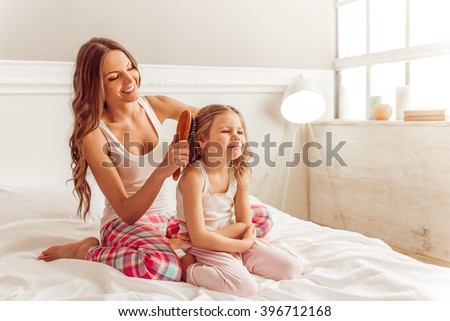 Beautiful young mother is combing her cute little daughter hair. Both in pajamas sitting on bed and smiling