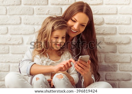 Beautiful young mother and her cute little daughter are listening to music using a smart phone and smiling, against white brick wall