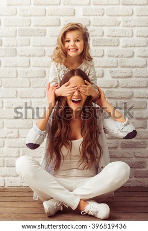 Young mother and her little daughter are looking at camera and smiling, sitting against white brick wall. Girl is closing her mother eyes