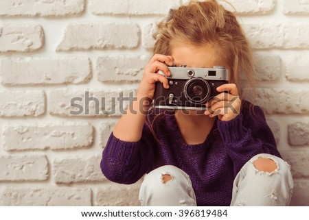 Beautiful little girl in casual clothes is taking a photo using a camera, looking at camera and smiling, sitting against white brick wall