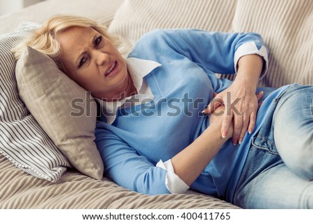 Beautiful tired old woman in casual clothes is having a stomach ache while lying on couch at home