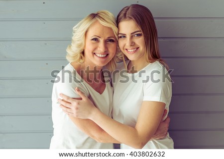 Portrait of beautiful mature mother and her daughter hugging, looking at camera and smiling, against gray background