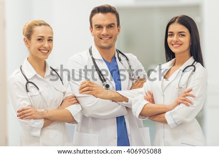 Three beautiful young doctors are looking at camera and smiling, standing with crossed arms in hall of the hospital