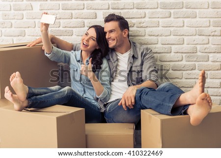 Happy attractive young couple is moving, making a selfie, cuddling and smiling while sitting among cardboard boxes