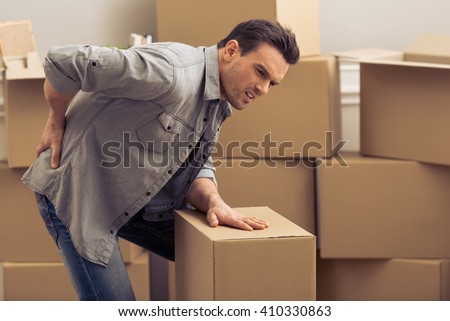 Handsome young man is moving, having pain in his back while packing, standing among cardboard boxes