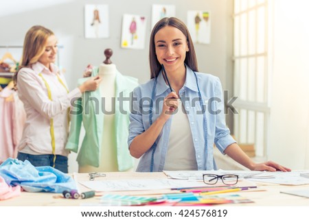 Beautiful designer is working in dressmaking studio, looking at camera and smiling, in the background another designer is working with jacket