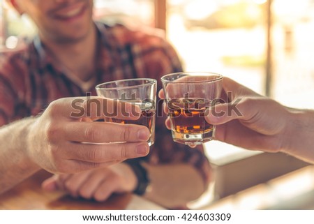 Cropped image of two men clanging glasses of alcoholic beverage together while sitting at bar counter in a modern urban cafe