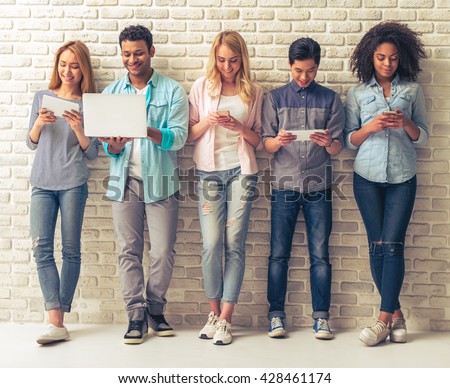 Beautiful young people of different nationalities are using gadgets and smiling, standing against white brick wall