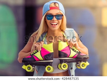 Portrait of beautiful blonde girl in cap and glasses looking at camera and smiling while standing with her rollers in skate park