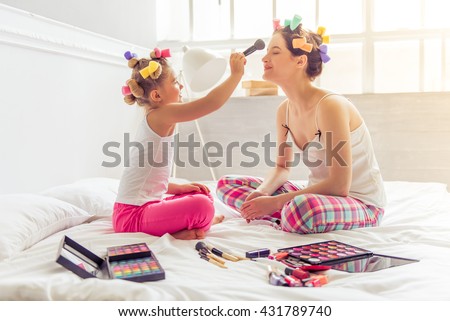 Beautiful young mother and her daughter in pajamas and with hair curlers on bed at home. Cute little girl is doing her mother makeup