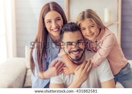 Portrait of beautiful young mother, father and their daughter hugging, looking at camera and smiling while sitting on sofa at home
