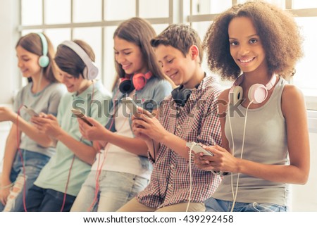Attractive teenage Afro American girl with headphones is using a smartphone, looking at camera and smiling while sitting among other teenagers