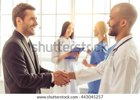 Handsome businessman and Afro American doctor are shaking their hands and smiling, two female doctors are talking in the background