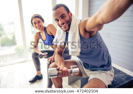 Beautiful Afro American couple in sports clothes is making selfie, holding dumbbells, looking at camera and smiling while resting after workout