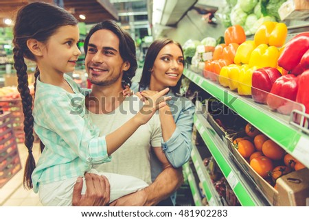 Beautiful young parents and their cute little daughter are talking and smiling while choosing food in the supermarket. Girl is sitting on dad's arms